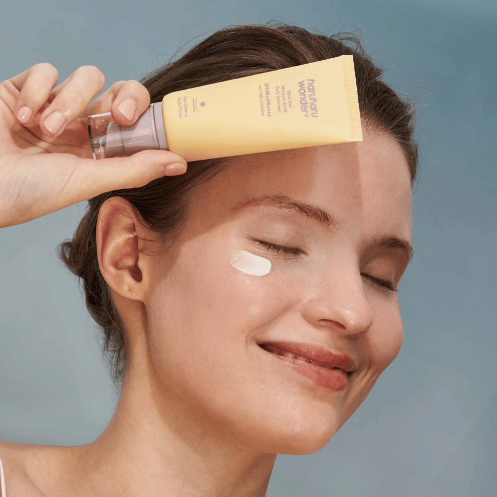 En leende kvinna applicerar Black Rice Moisture Airyfit Daily Sunscreen SPF 50+ PA++++ från Haruharu Wonder på sitt ansikte. Hon håller en gul tub med solkräm och har en klick av produkten på kinden. Bakgrunden är ljusblå.
