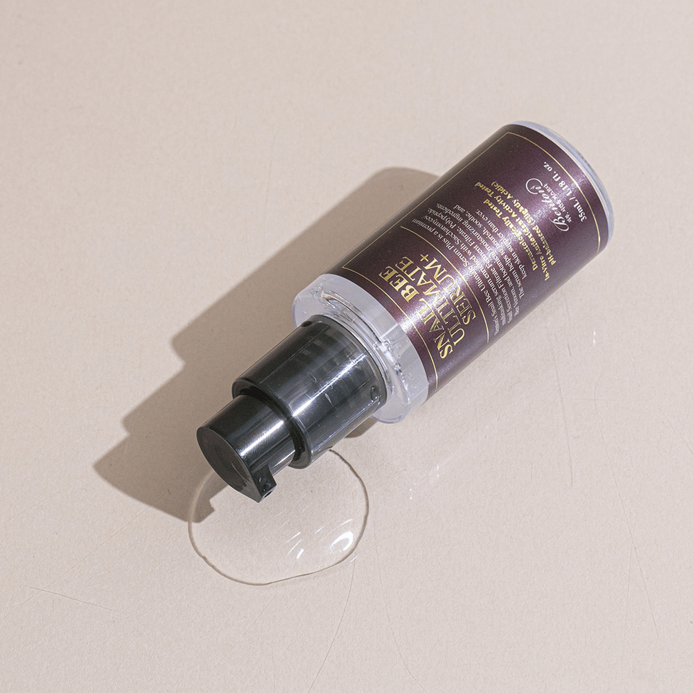 En flaska Snail Bee Ultimate Serum Plus ligger på en ljus yta med en liten pöl av vätska bredvid. Flaskan har en mörk etikett med guldtext och en svart pump.